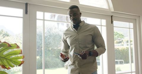 African American man standing by sunlit patio doors holding smartphone, wearing earbuds