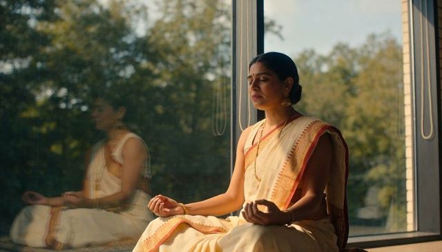 Indian woman meditating on sunlit window ledge with reflection wearing cream saree