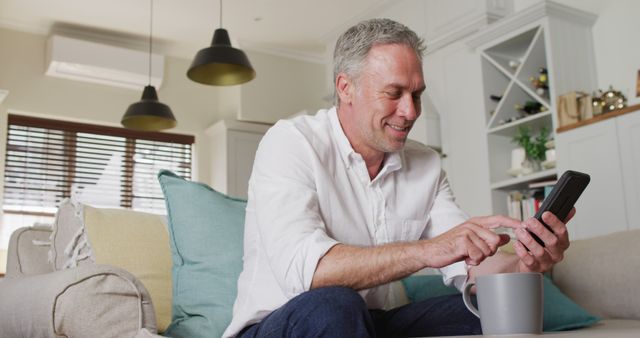 Mature man with grey hair sitting on a sofa in a modern living room, smiling while using a smartphone. The setting includes a contemporary design with soft cushions and modern decor. Perfect for promoting digital communication, lifestyle blogs, technology use among older adults, or home decor inspiration.