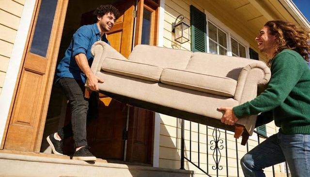 Smiling friends carrying beige sofa down porch steps during sunny home move