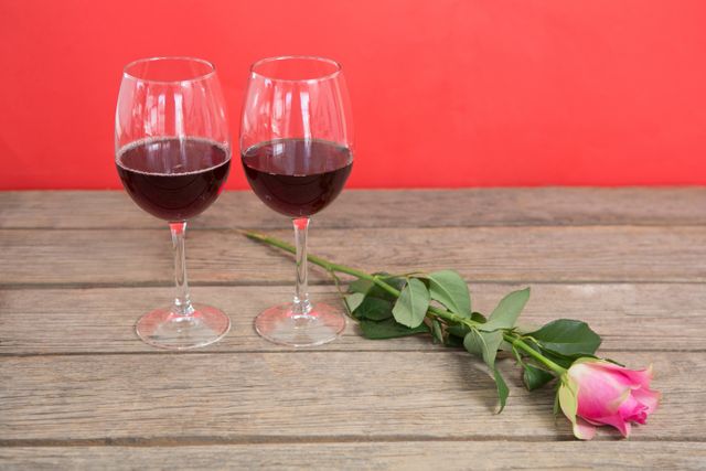 Two red wine glasses and a pink rose on a wooden surface with a red background. Ideal for romantic themes, Valentine's Day promotions, anniversary celebrations, and date night advertisements. Perfect for use in greeting cards, social media posts, and marketing materials related to love and romance.