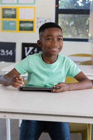 African american schoolboy smiling and using tablet with stylus in bright classroom