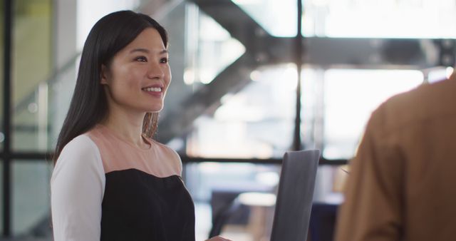 Asian businesswoman is smiling and engaging in conversation with a colleague in a modern office. Likely used for themes related to business communication, professional interactions, teamwork, corporate environments, and contemporary work settings.