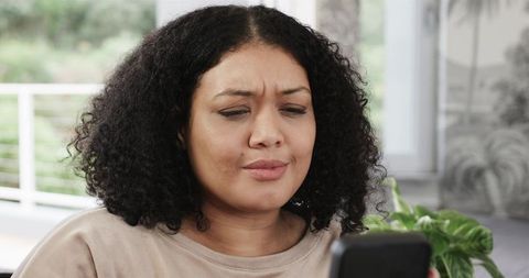 Young woman frowning at smartphone reacting to message while sitting in sunlit lounge