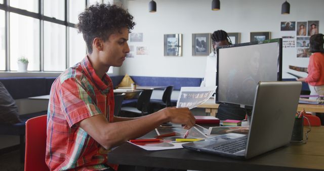 Young male designer working on a project using computer and printed materials in a modern office. He is wearing casual clothes and the office environment looks contemporary with large windows providing natural light. This can be used for presentations, websites, or articles related to creativity, design processes, modern work environments, or teamwork.