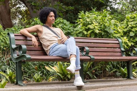 African american man relaxing on bench holding to-go coffee cup, casual park moment
