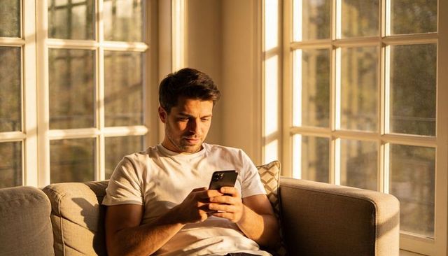 Young man checking smartphone on sunlit sofa near window during golden hour