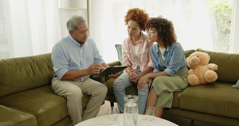 Senior father reviewing tablet with adult daughters on sofa, multigenerational family discussion