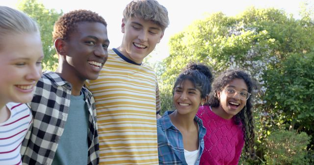 The image shows a diverse group of teenage friends standing in a line outdoors, smiling and enjoying their time together. They appear to be bonding and having fun under the sun, surrounded by greenery. This image is perfect for promoting themes like diversity, friendship, youth community, and carefree summer activities. It can be used in advertising campaigns, brochures, blogs, or educational materials.
