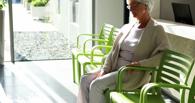 Elderly woman sitting on green chair in bright, modern medical office reception area with sunlight streaming in. Useful for depicting healthcare environments, senior patient experiences, or waiting areas in medical facilities.