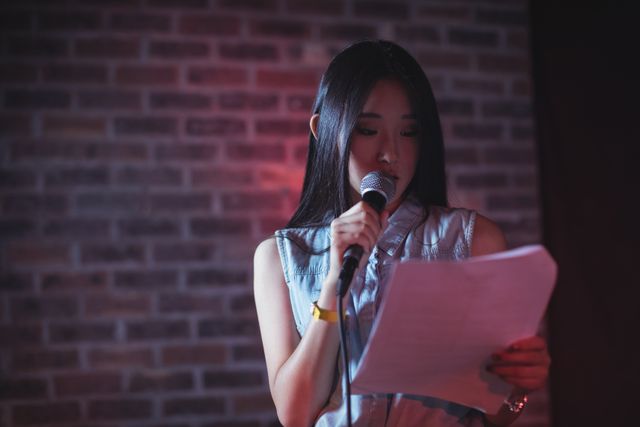 Young woman reading from a paper while holding a microphone on stage in a nightclub. Ideal for use in articles or advertisements related to public speaking, entertainment, nightlife, confidence, and young adults. Can also be used for promoting events, performances, and presentations.