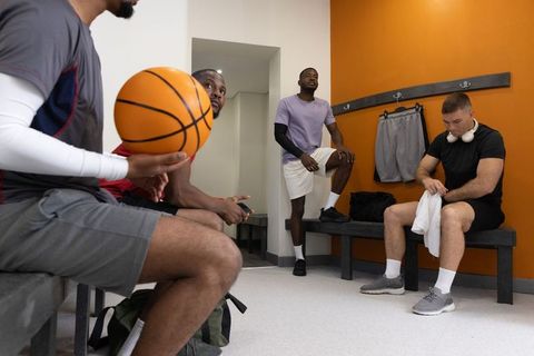 Multiethnic teammates sitting in locker-room holding basketball and preparing for game
