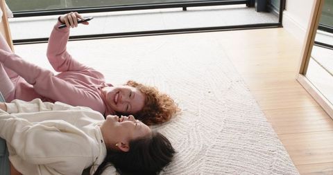 Multicultural friends lying on rug laughing while holding smartphone near sliding glass door