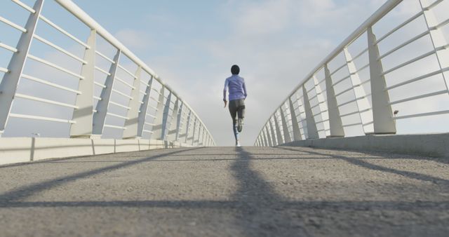 Rear view of biracial woman wearing hijab and sports clothes running on bridge in city. City living, fitness and healthy modern urban lifestyle.