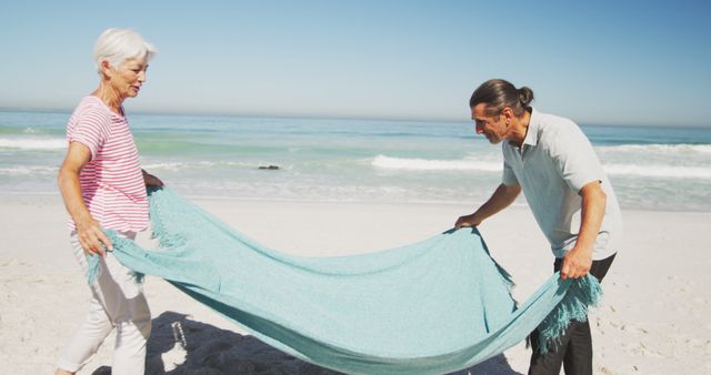 Senior couple spreading blue blanket on sandy beach by the ocean. Suitable for content involving retirement, travel, leisure activities, and elderly lifestyle. Ideal for advertisements promoting beach vacations, senior living, or outdoor recreation for older adults.