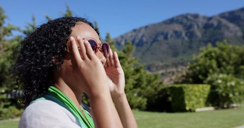 Adjusting round sunglasses in sunlit garden with green scarf and mountain backdrop