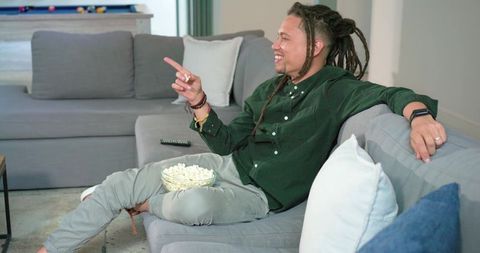 Young man relaxing on modern gray sofa enjoying popcorn and pointing at tv in lounge