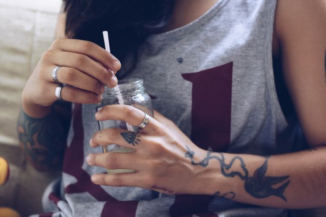 Young woman with tattoos and rings holds a drink in a mason jar with a straw. Casual indoor setting, ideal for concepts related to modern lifestyle, fashion, personal expression, and relaxation.