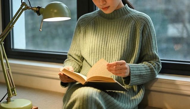 Asian woman reading on window seat wearing green sweater dress under lamp light