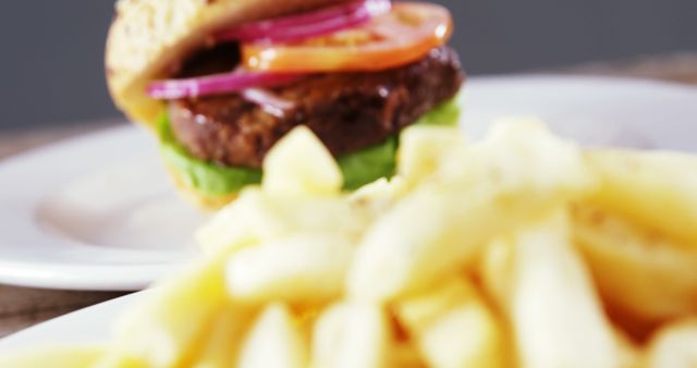 A close-up of a plate with French fries in focus, with a blurry hamburger in the background, with copy space. The image captures a common fast-food meal, emphasizing the texture and appeal of the fries.