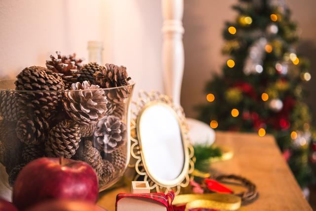 Christmas decorations on a wooden table including pinecones, gifts, apples, and a small mirror. A decorated Christmas tree with lights is visible in the background. Ideal for holiday-themed content, home decor inspiration, festive season promotions, and celebration advertisements.