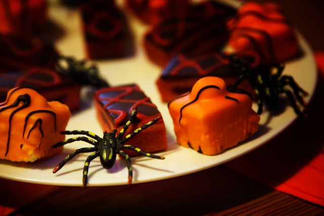 Halloween-themed dessert table featuring an assortment of sweets decorated with orange icing and chocolate lines, accompanied by toy spiders. Suitable for marketing materials during the Halloween season or festive event invitations and decorations.