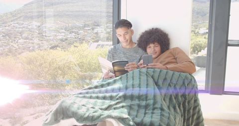 Cuddling on sunlit window seat reading book and checking phone under cozy blanket