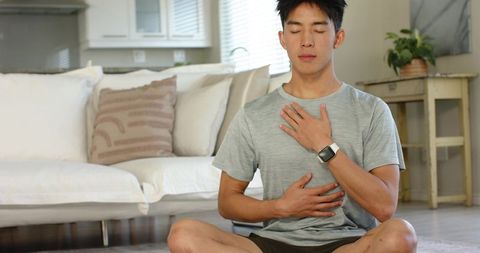 Asian man meditating in living room with smartwatch practicing mindful deep breathing