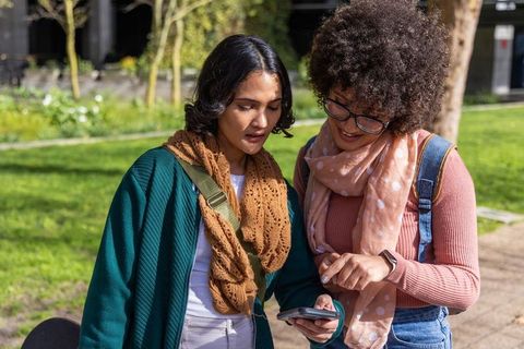 Diverse female students checking smartphone on campus path, pointing and laughing together