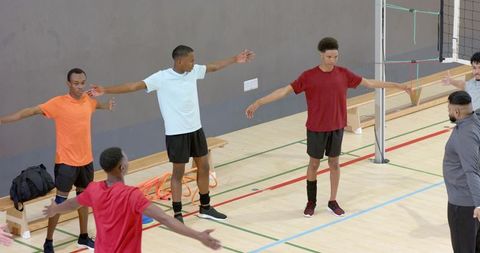 Diverse male volleyball team warming up with coach in gymnasium