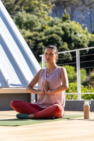 Woman practicing rooftop yoga sitting cross-legged on green mat wearing tank top at sunrise
