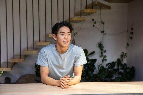 Young asian man leaning on light wood counter near floating staircase and indoor plants