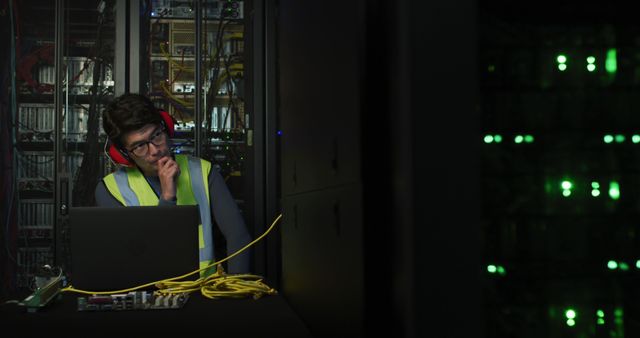 Male IT engineer working during nighttime in data center server room. Man wearing high-visibility vest, using laptop and monitoring equipment. Suitable for illustrating IT infrastructure, 24/7 operations, network engineering, and tech profession scenes.