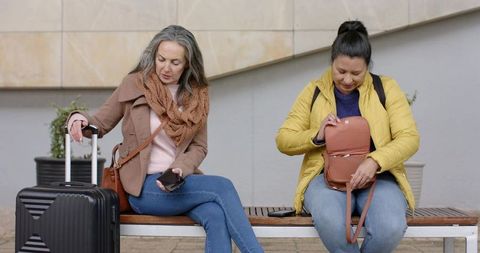 Mature women waiting on bench with suitcase and backpack in urban plaza during travel day