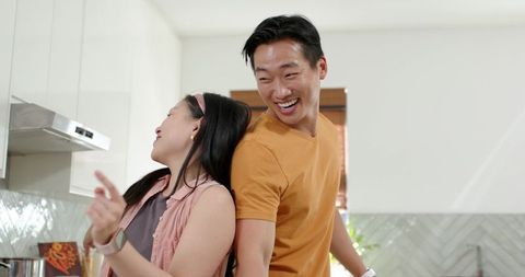 Asian couple laughing and leaning back-to-back in sunlit modern kitchen