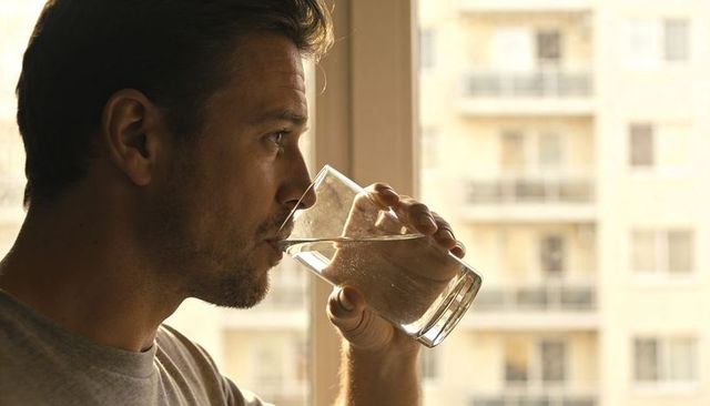 Man Drinking Water by Apartment Window with Urban Balcony View, Backlit Morning Light
