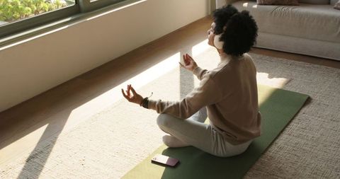 African american man meditating on yoga mat by sunlit window wearing headphones in loungewear
