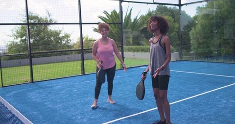 African american women playing padel on blue court, chatting and holding paddles by net