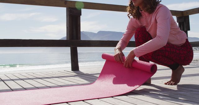 This image shows a woman in casual fitness clothing unrolling a pink yoga mat on a beachfront boardwalk. The scene implies an outdoor yoga session with a stunning ocean and mountain backdrop. Ideal for topics related to fitness, well-being, outdoor exercise, beach yoga retreats, and active, healthy lifestyle promotions.