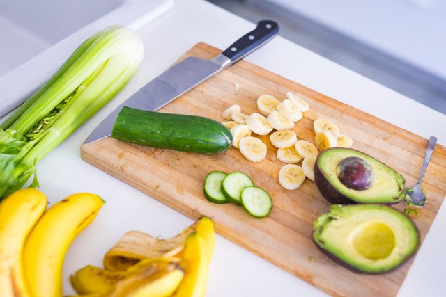 High angle view of fresh fruits and vegetables on a cutting board in a kitchen. Includes avocado, banana, cucumber, and celery. Ideal for use in articles or advertisements about healthy eating, vegan or vegetarian diets, home cooking, and organic food preparation.