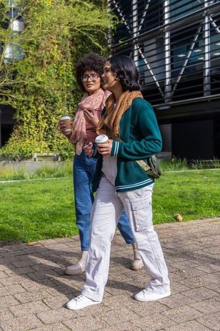 Two friends strolling with takeaway coffee along urban green wall near modern building