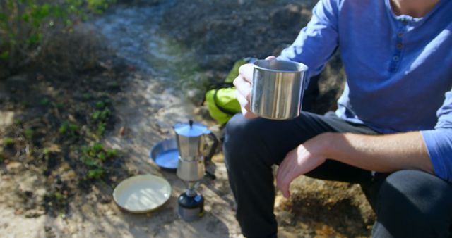 Hiker sitting in natural setting, holding a metal cup and enjoying a hot beverage. Ideal for travel blogs, outdoor adventure magazines, and promoting camping gear. Highlights the simplicity and relaxation of camping life.