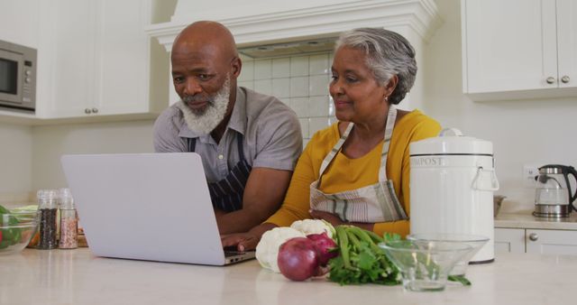 Senior African American couple in kitchen cooking dinner, using laptop for recipe while surrounded by fresh vegetables like onions, lettuce and cauliflower. They are smiling and seem to be enjoying their time together. Useful for content related to technology use among seniors, healthy eating, elderly lifestyle, bonding activities and multicultural cooking.