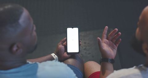 Two african men checking fitness app on smartphone while sitting side-by-side wearing wrist trackers
