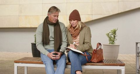 Mature couple sitting on bench in urban plaza, woman writing in journal, cozy fall outfits