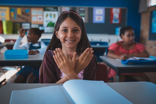 Portrait of smiling caucasian schoolgirl in classroom sitting at desk making bird shape with hands - Download Free Stock Photos Pikwizard.com