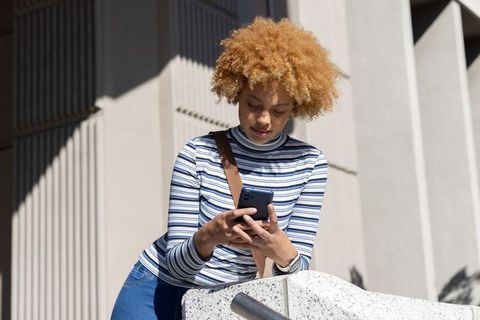 African american woman checking smartphone on urban concrete ledge in sunlight