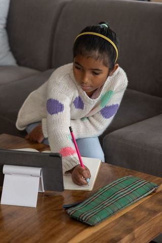 Indian girl studying at home writing in notebook while following tablet lesson on sofa