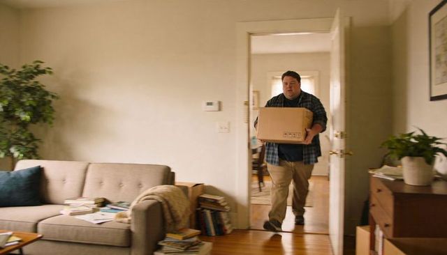 Man carrying sealed moving box into cozy living room with sofa, plants and stacked books