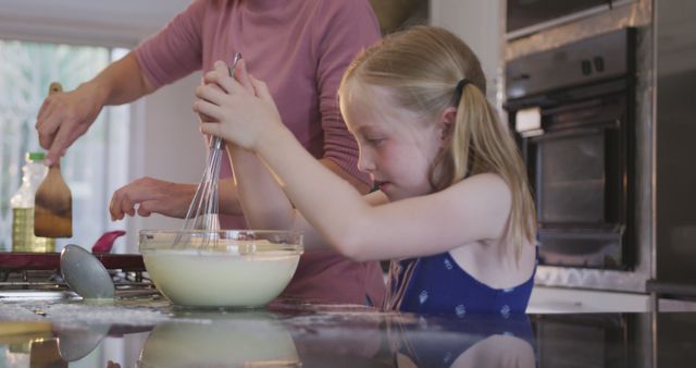 Young girl in blue dress mixing batter with a whisk while a parent cooks at the stove. Ideal for images depicting family activities, cooking lessons, or domestic life. Can be used for blogs, social media, parenting magazines, and advertisements related to home cooking and family bonding.